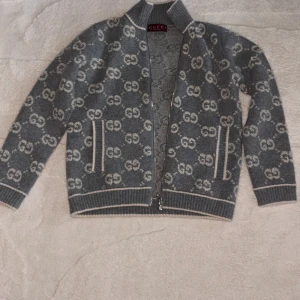 Grå stickad sweater från Gucci - Säljer en grå stickad sweater från Gucci med ikoniskt GG-mönster över hela jackan. Sweatern har dragkedja framtill, ribbade muddar och krage samt vita detaljer vid fickor och kanter. Perfekt för dig som vill ha en lyxig och trendig look. Helt ny inte använd en ända gång finns även lappar samt påse!