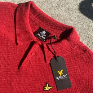 Röd pikétröja från Lyle & Scott - Röd pikétröja från Lyle & Scott med klassisk krage, tre knappar och det ikoniska gula fågel-loggan på bröstet. Tröjan är stickad i bomull och har långa ärmar samt ribbade muddar och nederkant. Perfekt för dig som gillar stilren och sportig look.