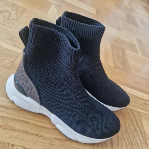 Svarta stickade sneakers Michael Kors - Snygga svarta stickade sneakers från Michael Kors med högt skaft och vit chunky sula. Baksidan har bruna detaljer med MK-logga i mönster. Skorna är i stretchigt syntetmaterial och har en sportig, modern look som sticker ut.