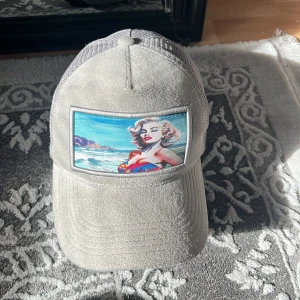 Grå meshkeps från Revenite med tryck (pr kan diskuteras) - Grå keps från Revenite med mesh på sidorna och baksidan. Framtill finns ett färgglatt tryck med strandmotiv och retroinspirerad illustration. Kepsen har böjd skärm och justerbar passform. Perfekt för dig som vill sticka ut med en unik accessoar.