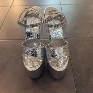 Silvriga platåsandaler med 17cm hög klack - Säljer ett par riktigt snygga silvriga sandaler från Binsheng med 17cm höga klackar. Skorna har ett glansigt, spräckligt mönster och smala remmar med guldfärgade spännen. Perfekta för dig som vill sticka ut och älskar statementskor.