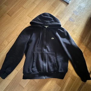 Svart Lacoste zip - Är i helt nytt skick! Är dock använd typ 1-2 gånger. Modellen på bilden är 175cm och väger 71kg