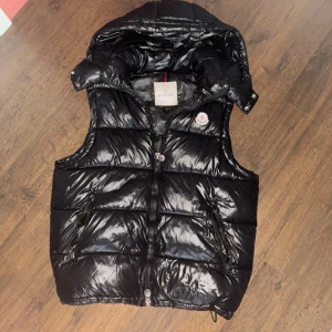 Svart dunväst från Moncler med huva - Svart glansig dunväst från Moncler med avtagbar huva och dragkedja framtill. Västen har två fickor med dragkedja och klassisk Moncler-logga på bröstet. Perfekt för lager-på-lager och streetstyle. Fodrad med dun och har justerbar dragsko nertill.
