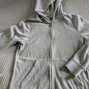 Grå Juicy Couture hoodie i velour - Supermjuk grå hoodie från Juicy Couture i velour med dragkedja och huva. Ikonisk logga broderad i guld på ryggen och snygg J-detalj på dragkedjan. Perfekt för en chill och trendig look. Strl M men är för liten för mig som är 38, så jag skulle rekommendera en strl 36 att köpa. Då kommer den sitta perfekt så som "den ska".