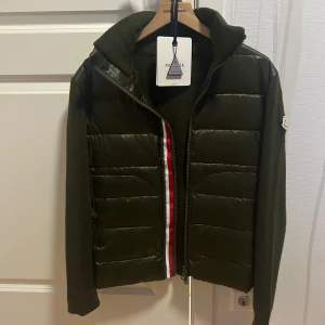 Moncler Cardigan  - Snygg olivgrön pufferjacka från Moncler med stickade ärmar och rygg. Jackan har quiltad framsida i glansigt material, dragkedja med röd, vit och marinblå detalj samt hög krage. Perfekt för dig som vill ha en stilren och sportig look. 