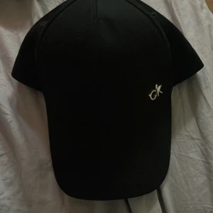Svart keps från Calvin Klein - Svart keps från Calvin Klein med klassisk böjd skärm och justerbart spänne bak. Framtill finns en liten silverfärgad 'ck'-logga och på sidan syns Calvin Klein-tryck i vitt. Snygg och stilren accessoar som passar till allt.