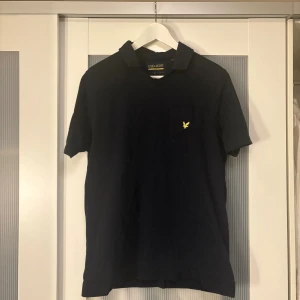 Mörkblå t-shirt från Lyle & Scott - Snygg mörkblå t-shirt från Lyle & Scott med klassisk krage och liten bröstficka med gul logga. Tillverkad i mjuk bomull, perfekt för en clean och stilren look. Passar dig som gillar enkel men ikonisk design. 