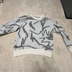 Gråvit stickad tröja från Manfinity - Säljer en stickad tröja från Manfinity i storlek M. Tröjan är gråvit med abstrakt mönster i mörkgrått och har ribbade muddar vid ärmslut, hals och nederkant. Perfekt för dig som gillar en chill och modern stil.