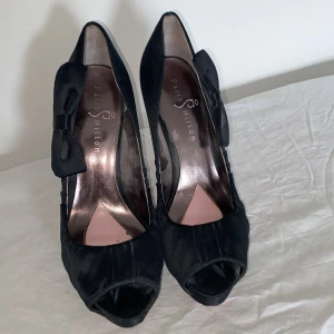 Svarta pumps från Paris Hilton - Snygga svarta pumps från Paris Hilton i satin med öppen tå och hög klack. Skorna har en elegant rosett på sidan och en cool rosa sula som sticker ut. Perfekta för dig som vill ha en klassisk men ändå trendig look.