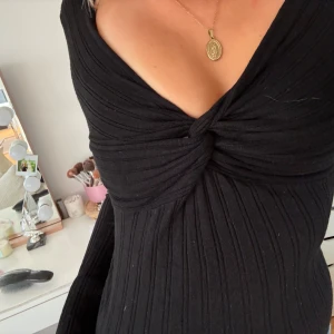 Svart ribbad långärmad topp med twist - Säljer en svart långärmad topp med ribbat tyg och snygg twistdetalj framtill vid bysten. Djup v-ringning ger en trendig look och passformen är tight. Perfekt att styla med jeans eller kjol för en cool vibe.