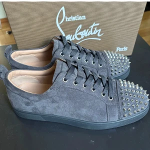 Christian Louboutin  - Mörkgråa sneakers i mocka från Christian Louboutin med ikonisk röd sula och silverfärgade nitar på tån. Skorna har snörning och rund tå, samt lyxig läderinsida. Perfekt för dig som vill sticka ut med en edgy och exklusiv look. Skriv om det är något ni undrar. Boxen och påsen till skorna samt og fås med vid köp. Priset går att diskutera under snabb affär