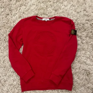 Röd Stone Island sweatshirt XL - Riktigt snygg röd sweatshirt från Stone Island i slim fit. Tröjan har broderad logga framtill och klassisk patch på ärmen. Mjuk bomullskvalitet och rund halsringning. Perfekt för dig som vill ha en stilren och trendig look. Den är i xl men passar S