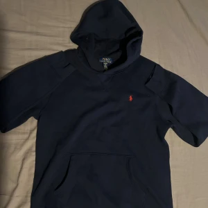 Mörkblå hoodie från Polo Ralph Lauren - Mörkblå hoodie från Polo Ralph Lauren med korta ärmar och klassisk huva. Tröjan har en röd broderad logga på bröstet och en stor magficka framtill. Perfekt för en chill och sportig look.