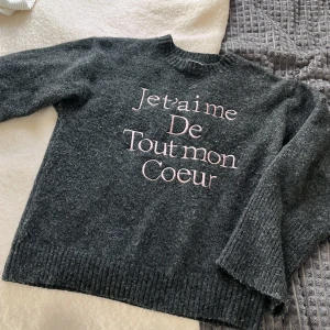 Mörkgrå stickad tröja text  - Mysig mörkgrå stickad tröja med rosa broderad text 'Je t'aime De Tout mon Coeur' på framsidan. Tröjan har rund halsringning, långa ärmar och ribbade muddar. Perfekt för kyliga dagar och ger en cool, avslappnad vibe.