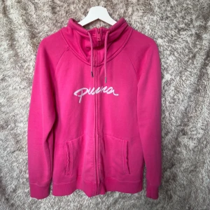 Rosa hoodie från Puma med dragkedja - Säljer en snygg rosa hoodie från Puma med vit broderad logga på bröstet och ärmen. Tröjan har en hög krage med snörning, två fickor fram och hel dragkedja. 