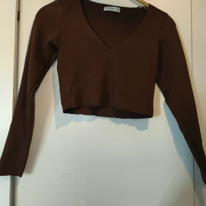 Brun croppad topp från Stradivarius - Snygg brun croppad topp från Stradivarius med lång ärm och djup v-ringning. Toppens material är ribbad och stretchigt, vilket gör den bekväm och följsam. Perfekt att styla med höga jeans eller kjol för en trendig look.