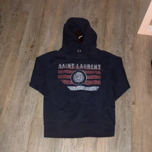 Saint Laurent hoodie - Mörkblå hoodie från Saint Laurent med tryck i vitt och rött framtill. Tröjan har huva med snörning och klassisk känguruficka. Perfekt för dig som gillar en avslappnad stil och vill ha en snygg logga på bröstet. Är S men sitter som XS              Priset kan diskuteras vid snabb affär 