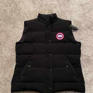Svart dunväst från Canada Goose - Svart dunväst från Canada Goose med klassisk logga på bröstet. Västen har hög krage, tryckknappar framtill och två snedställda fickor. Perfekt för lager-på-lager och riktigt snygg till streetwear eller outdoor-stil.