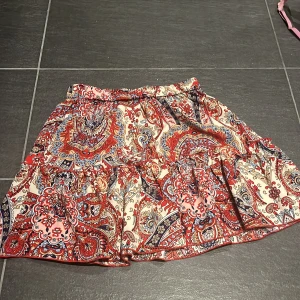 Mönstrad kortkjol med volang - Supersöt kortkjol i rött, blått och beige. Kjolen har volangdetaljer och resår i midjan för en bekväm passform. Kan även användas som en fin topp🩷