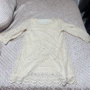Beige spetsklänning med trekvartsärm - Superfin beige spetsklänning med vackra detaljer och trekvartsärmar. Klänningen har en rund halsringning och ett fint mönster i spets över hela plagget samt dekorativ kant nertill. Perfekt för dig som gillar romantisk och feminin stil.