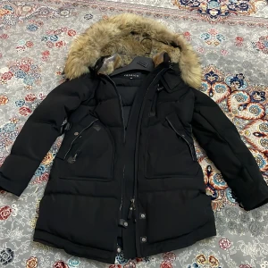 Svart dunjacka med fuskpäls från Cedrico - Säljer en svart dunjacka från Cedrico i storlek XS. Jackan har en stor huva med mjuk fuskpäls, dragkedja och knappar framtill samt flera praktiska fickor. Perfekt för kalla dagar med sin långa modell och vadderade design. Köpte den för 8000kr säljer för billigare. 