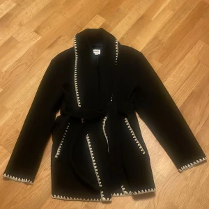 Svart omlottkappa med vita detaljer - Svart kappa från Kappahl med omlottmodell och bälte i midjan. Jackan har vita sicksack-detaljer längs kanterna och ärmarna, samt två fickor framtill. Stilig och trendig design som sticker ut.