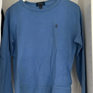 Blå långärmad tröja Polo Ralph Lauren - Blå långärmad tröja från Polo Ralph Lauren med klassisk rund halsringning och broderad logga på bröstet. Tillverkad i mjuk bomull, perfekt för en avslappnad och stilren look.