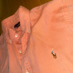 Rosa skjorta från Ralph Lauren S - Snygg rosa skjorta från Ralph Lauren i slim fit med klassisk button down-krage och broderad logga på bröstet. Skjortan är i bomull och har långa ärmar samt vita knappar framtill. 