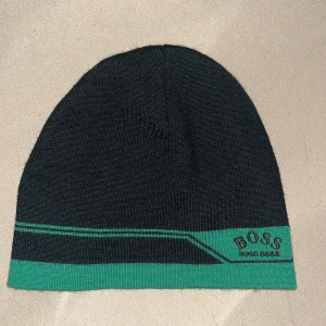 Svart och grön mössa från BOSS - Snygg svart mössa från BOSS med grön kant och diskret logga framtill. Mössan har en enkel, tight passform och är tillverkad i mjukt stickat material. Perfekt för dig som vill ha en stilren accessoar med en sportig touch.