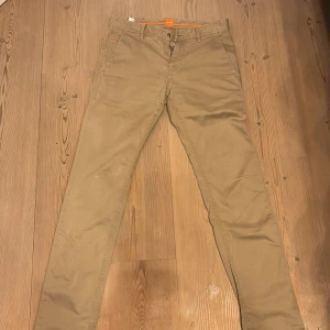Beiga chinos från BOSS slim fit - Snygga beiga chinos från BOSS i slim fit-modell. Byxorna har klassisk design med fickor fram och bak, bälteshällor och dragkedja. Tillverkade i mjuk bomull som ger en skön känsla och stilren look. Perfekta för dig som gillar en smalare passform. Varan saknar en knapp vid gylfen. Detta går dock enkelt att fixa.