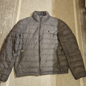 Grå pufferjacka från Polo Ralph Lauren - Grå pufferjacka från Polo Ralph Lauren med diskret logga på bröstet. Jackan har ståkrage, två dragkedjefickor och quiltad design. Tillverkad i syntetmaterial och har hel dragkedja framtill. Perfekt för kyliga dagar och enkel att matcha med det mesta. Har små defekter därav priset 