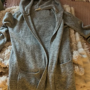 Grå kofta med huva från KappAhl - Mysig grå kofta från KappAhl med huva och två stora fickor framtill. Koftan är stickad och har omlott-design, perfekt för kyliga dagar. Långärmad och i en loose passform som ger en avslappnad vibe.