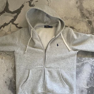 Grå hoodie från Polo Ralph Lauren - Snygg ljusgrå hoodie från Polo Ralph Lauren med dragkedja, huva med vita snören och klassisk blå logga på bröstet. Mjuk bomullskänsla och två stora fickor framtill. Perfekt för chill dagar eller när du vill ha en clean look.