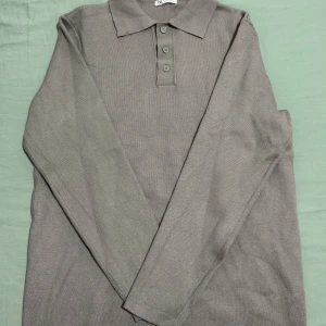 Grå långärmad pikétröja från Zara - Säljer en stilren grå pikétröja från Zara med klassisk krage och tre knappar framtill. Tröjan är långärmad och tillverkad i ett mjukt, stickat material som känns skönt mot huden. Perfekt för dig som gillar en clean och enkel look.