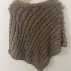 Brun pälsponcho med struktur - Säljer en brun poncho i mjuk päls med snedställda ränder som ger en cool struktur. Perfekt att slänga över en hoodie eller tröja för extra värme och stil. Den har en lös passform och är supermysig för kyliga dagar.