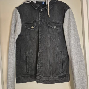 Svart jeansjacka med grå ärmar H&M Divided - Snygg svart jeansjacka från H&M Divided med grå sweatshirt-ärmar och huva. Jackan har två bröstfickor med knappar och två sidofickor. Perfekt för dig som gillar en avslappnad streetstil med mix av denim och mjukare material.