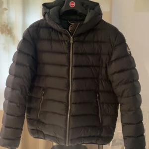 Svart dunjacka från Colmar - Säljer en svart dunjacka från Colmar med huva och dragkedja framtill. Jackan har quiltad design, två fickor med dragkedja och en logga på ärmen. Perfekt för kyliga dagar och riktigt snygg passform. Billigare vid snabb och smidig affär 