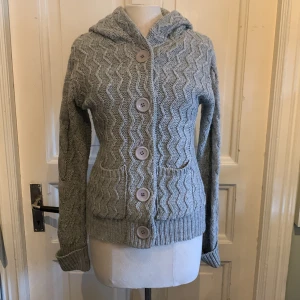 Grå stickad kofta med huva Capri Collection - Mysig grå stickad kofta från Capri Collection med huva och stora knappar framtill. Snyggt vågigt mönster, två fickor och ribbade muddar. Perfekt för lager-på-lager under kyliga dagar.