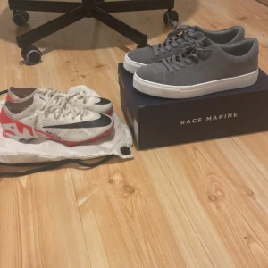 Grå Race Marine sneakers & Nike fotbollsskor - Säljer ett par stilrena grå sneakers från Race Marine med vit platt sula och snörning, samt ett par vita Nike fotbollsskor med svarta och röda detaljer, snörning och rund tå. Perfekt för dig som gillar både streetstyle och sport. Nike skorna ser kanske lite rustiga ut men kommer tvättas innan ni får den