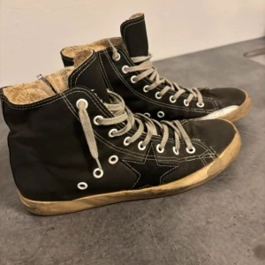 Golden goose  - Svarta höga sneakers med vit kontrastsöm och grå snörning. Sidan har en snygg stjärndetalj och sulan är beige med svarta undersidor. Skorna är tillverkade i canvas med insida i syntetmaterial. Klassisk streetstil som funkar till jeans och casual outfits.