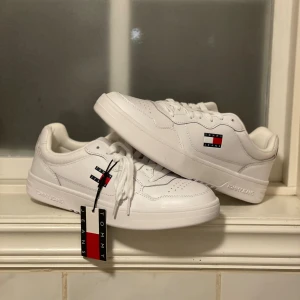 Vita sneakers Tommy Jeans - Fräscha vita sneakers från Tommy Jeans med klassisk låg siluett och rund tå. Skorna har snyggt broderad logotyp i rött, vitt och blått på sidan samt präglad text på sulan. Materialet är slätt syntet med snörning framtill för en clean look. Nypris 1200:- 