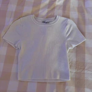 Vit ribbad topp från Zara - En stilren vit ribbad topp från Zara i storlek S. Toppen har rund halsringning, korta ärmar och en croppad passform. Perfekt att matcha med jeans eller kjol för en clean look.