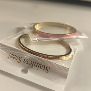 Guldiga armband med rosa detaljer - Två stilrena armband i guldton av stainless steel. Det ena har en rad små rosa stenar och det andra har en slät rosa emaljdetalj. Perfekta för att addera lite färg och glans till din look.