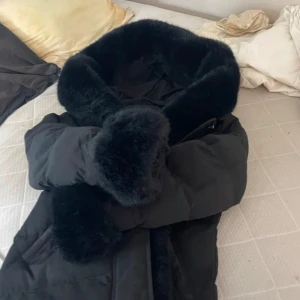 Svart pufferjacka med päls från Copperose - Säljer en svart pufferjacka från Copperose i storlek M. Jackan har en stor fluffig krage och muddar i pälsimitation som ger en lyxig vibe. Perfekt för kalla dagar när du vill vara både varm och snygg. Jackan har en klassisk pufferlook och är riktigt mysig.