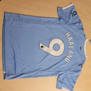 Manchester City Haaland #9 - Manchester Citys hemmatröja med Haaland och nummer 9 på ryggen. Tröjan är ljusblå med vita och mörkblå detaljer, har korta ärmar och är tillverkad i Drycell-material med minst 50% återvunnet material. Puma-logga och klubbmärke broderade på bröstet.