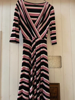 Randig långklänning från Jumperfabriken - Supersnygg långklänning från Jumperfabriken i svart, rosa och vit med coola diagonala och horisontella ränder. Klänningen har omlott-v-ringning och trekvartsärm. Mjuk och stretchig polyester som sitter skönt och ger en riktigt chic look.
