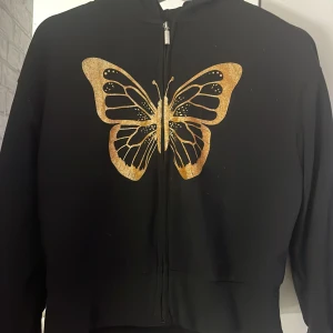 Svart hoodie med guldglittrig fjäril - Cool svart hoodie med dragkedja och huva, prydd med en stor guldglittrig fjäril på framsidan. 