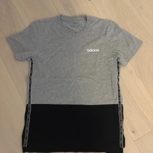 Grå och svart t-shirt från Adidas - Snygg t-shirt från Adidas i storlek S med grå överdel och svart nederdel. T-shirten har korta ärmar och rund hals. Längs sidorna finns svarta band med Adidas-logga. Materialet är mjuk bomull, perfekt för en chill och sportig look.