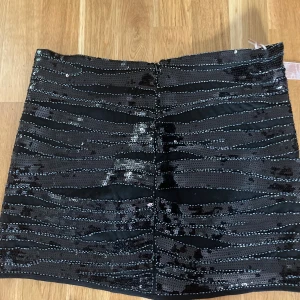 Svart paljettkjol från Sweewë - Själv skulle jag säga storlek M.Snygg svart kortkjol från Sweewë med glittriga paljetter i vågiga ränder. Kjolen har en figurnära passform och dragkedja bak. Perfekt för dig som vill sticka ut med en glammig look. Materialet är polyester och kjolen har en cool, festlig vibe.