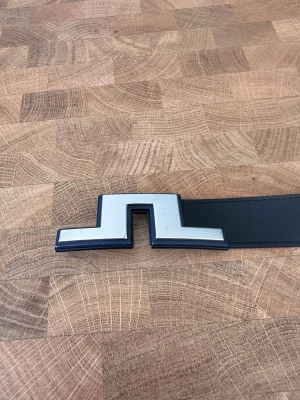 Svart skärp från J.Lindeberg i skinn - Snyggt svart bälte från J.Lindeberg i äkta skinn, tillverkat i Italien. Bältet har en unik, geometrisk silverfärgad spänne som ger en modern vibe. Perfekt för dig som vill ha en stilren accessoar med lite edge.